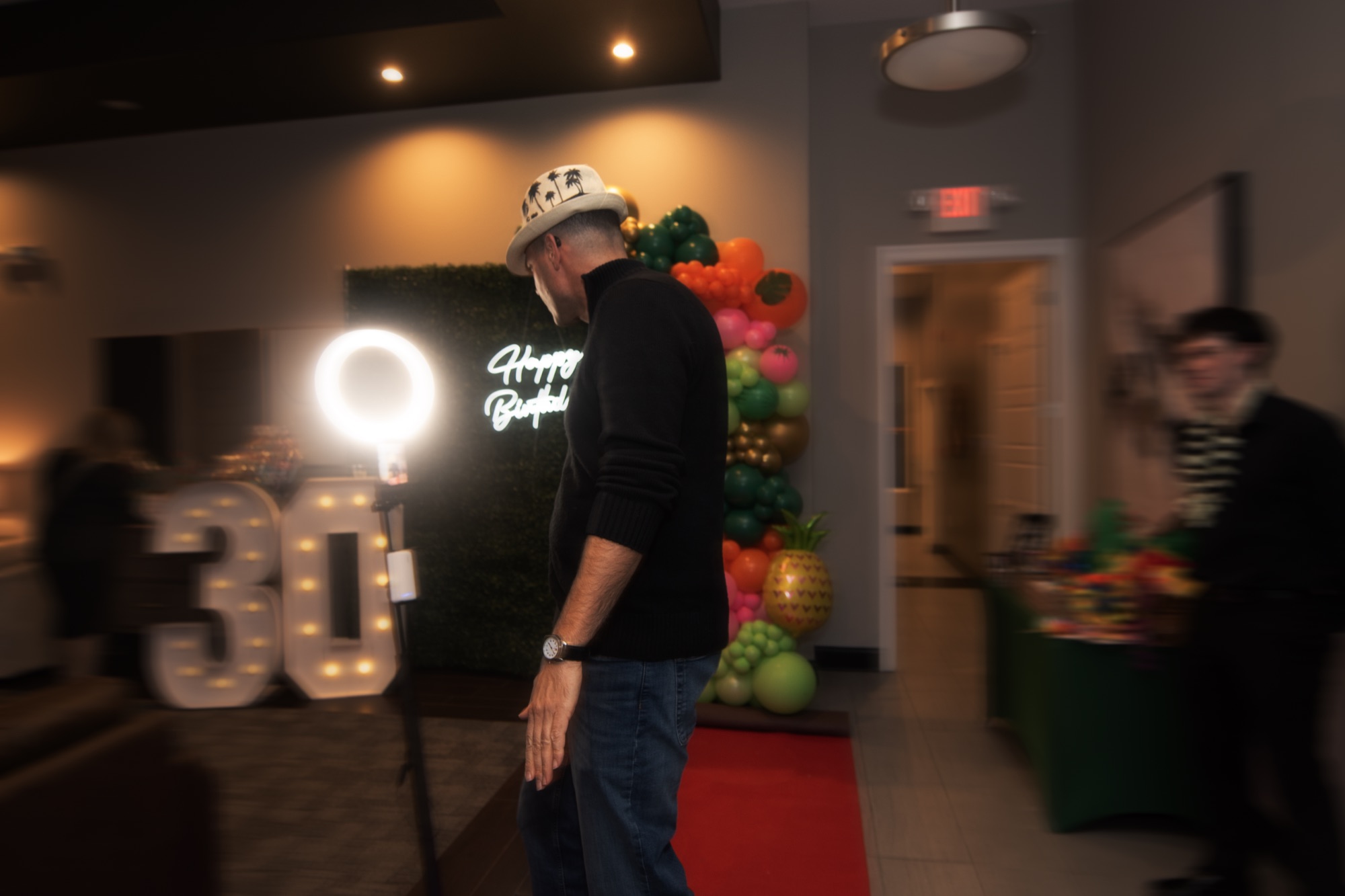 Guest wearing a fedora stepping toward the NextSpin 360 booth at a tropical-themed 30th birthday with marquee numbers and a balloon garland.